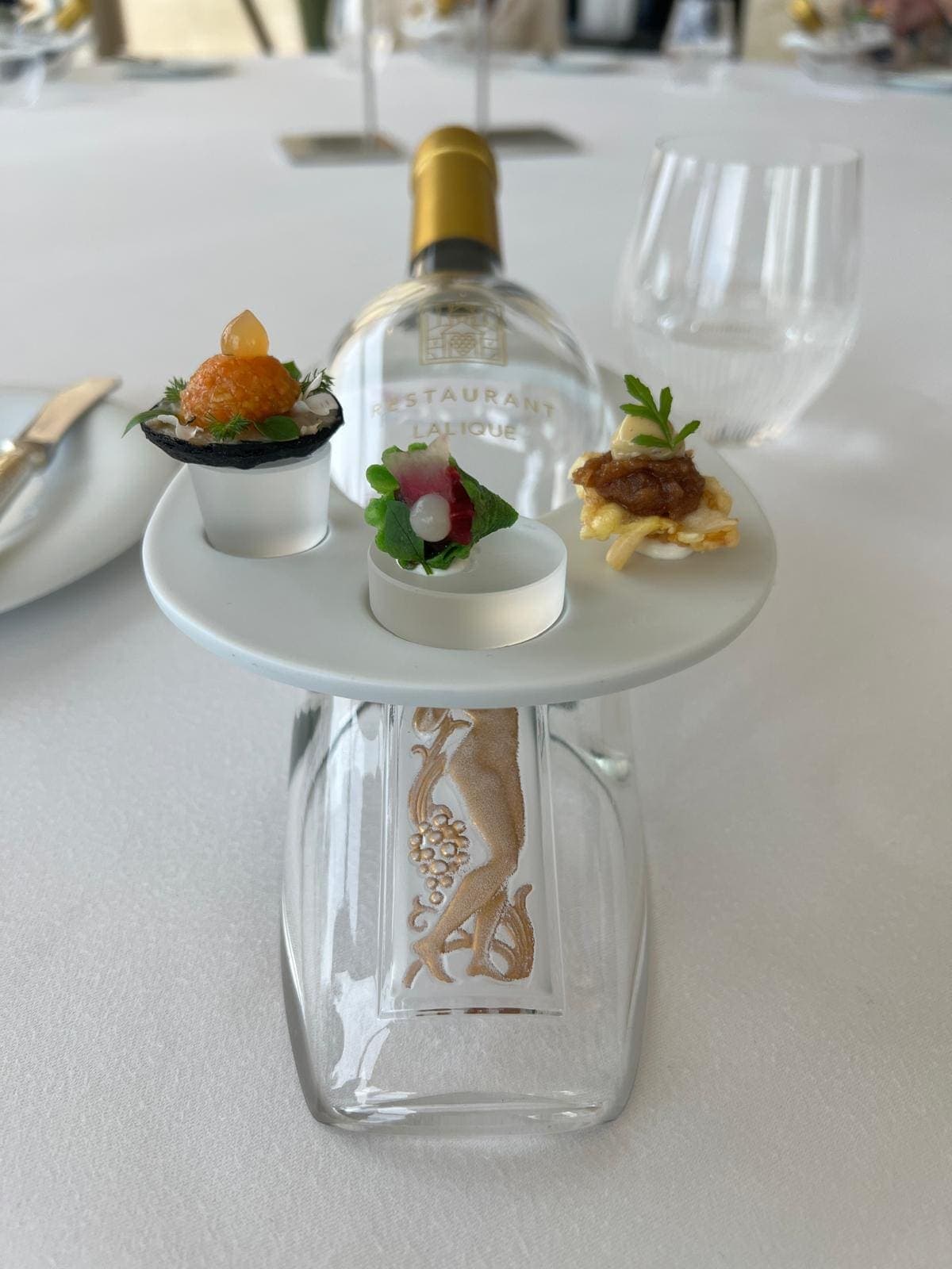 Présentoir de trois amuse-bouches sur un socle en verre gravé « Restaurant Lalique », bouteille d’eau en fond