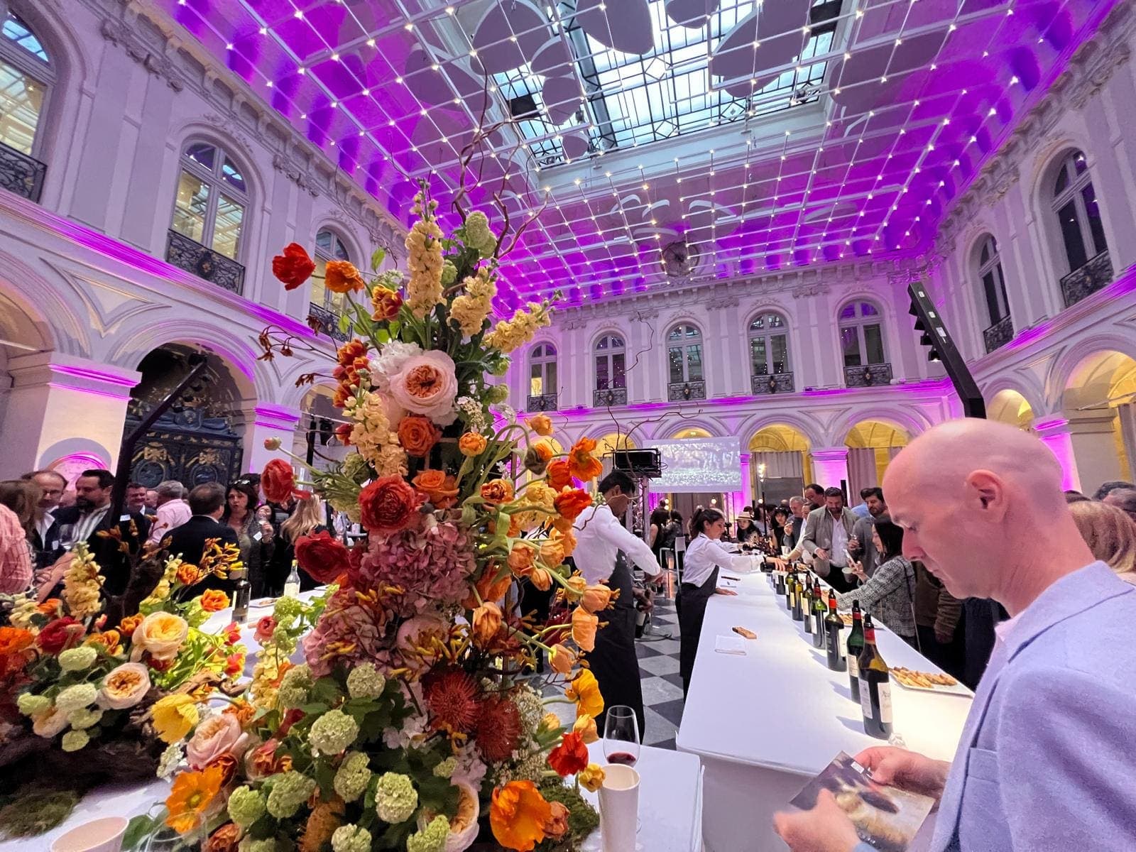 Réception dans une cour d’hôtel particulier à la verrière éclairée de pourpre, grand bouquet de fleurs au premier plan