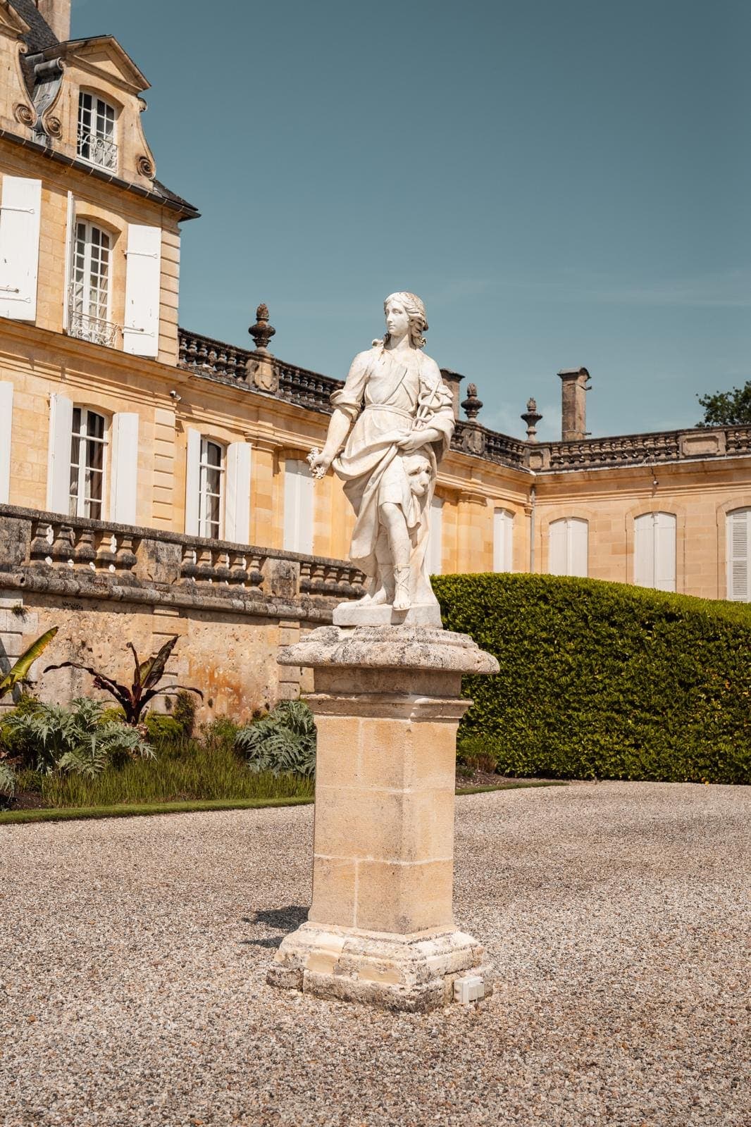 Statue en pierre sur un piédestal dans la cour gravillonnée d’un château, avec haie taillée