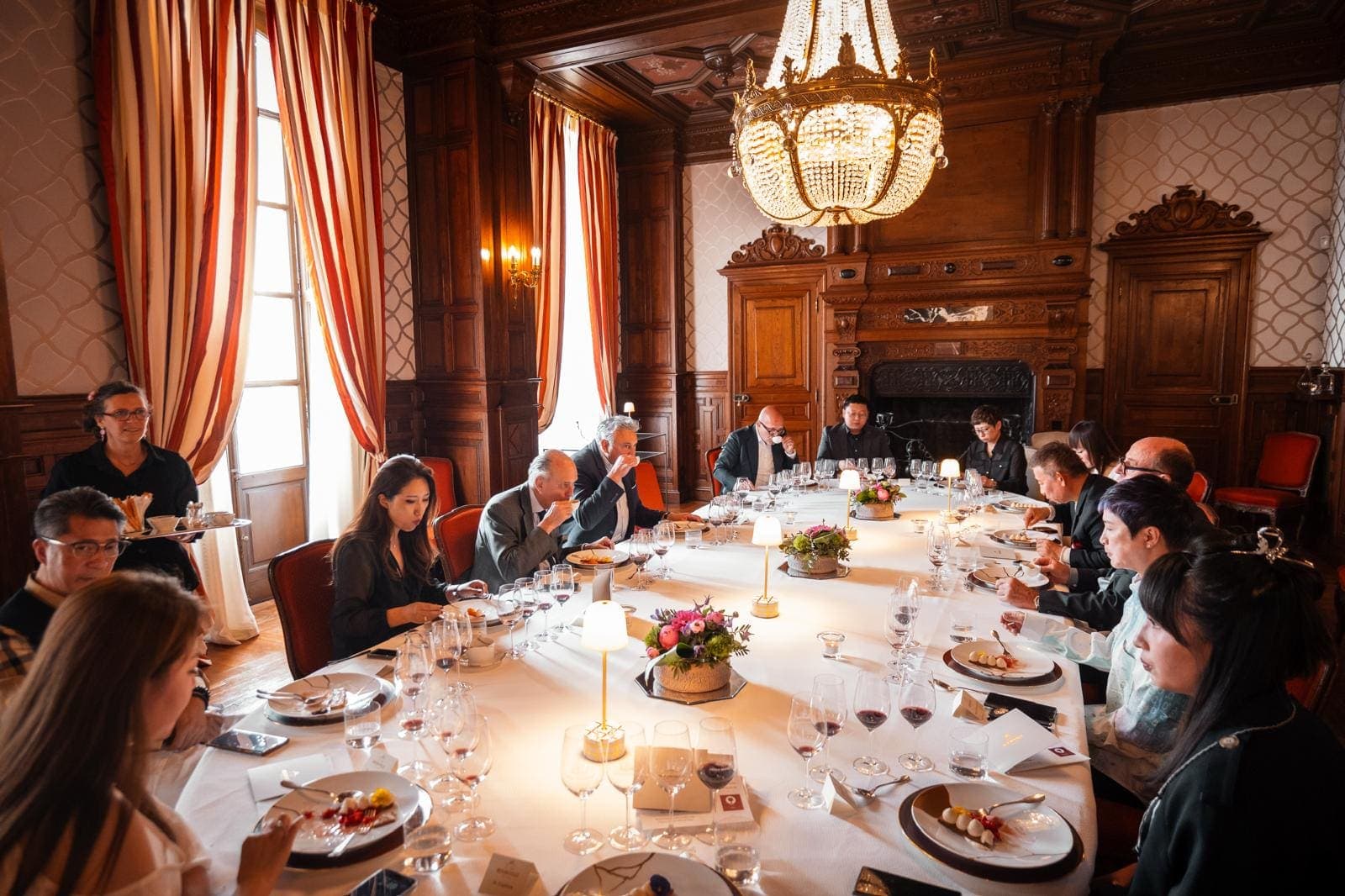 Longue table dressée sous un lustre en cristal, dans une salle à boiseries et rideaux rouges de château